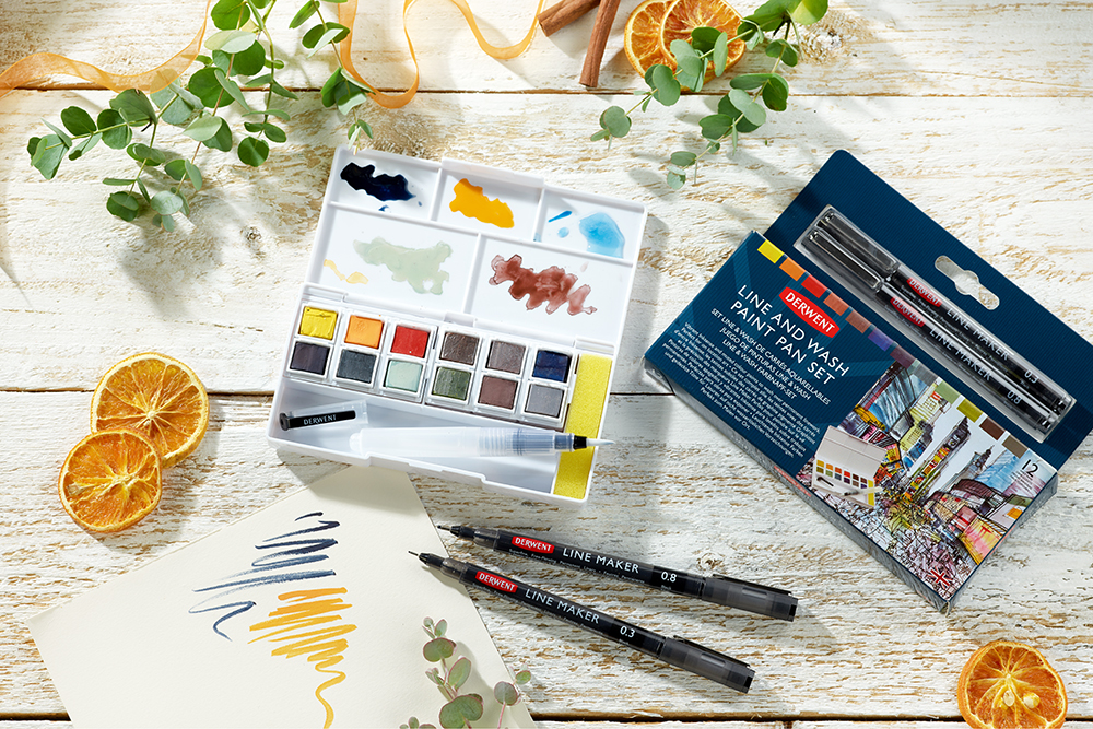 The Derwent Line and Wash Paint Paint Pan Set is photographed on a white wooden background. Near the set is the box and two Line Maker fine liner pens, as well as a sheet with swatches of colour.