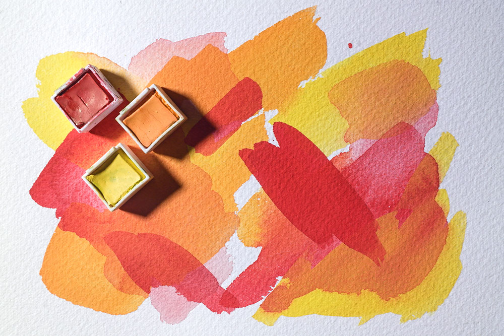 Inktense Pans of Sun Yellow, Mango and Poppy Red sit on a background painted with bright swatches of each coour. The colours are painted with different opacities and overlap without bleeding.