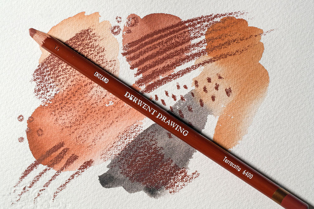 A Derwent Terracotta Drawing Pencil is photographed on a sheet of paper. The paper is covered with expressive pencil strokes in a rich brown colour. Paint has been layered over the pencil strokes.