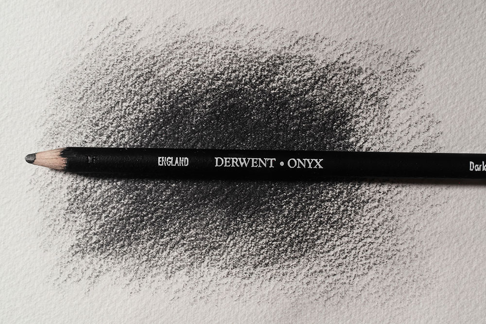 A Derwent Onyx pencil rests on a piece of paper covered in black strokes of pencil.