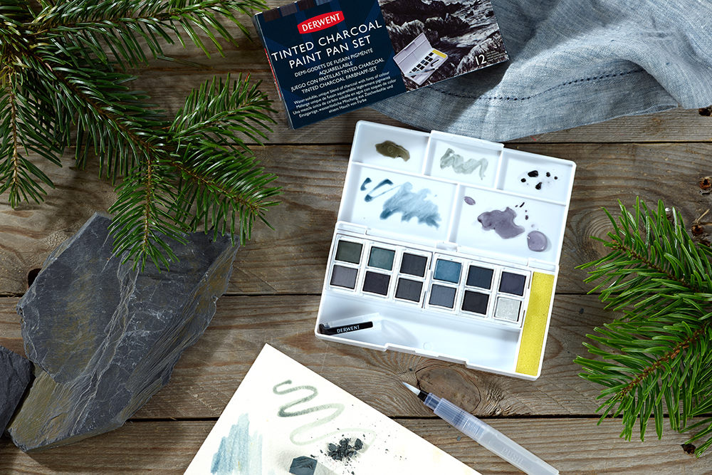 The Derwent Tinted Charcoal Paint Pan Set photographed on a wooden background. A paper sheet includes swatches of some colour and the set is surrounded with slate chunks and pine sprigs.