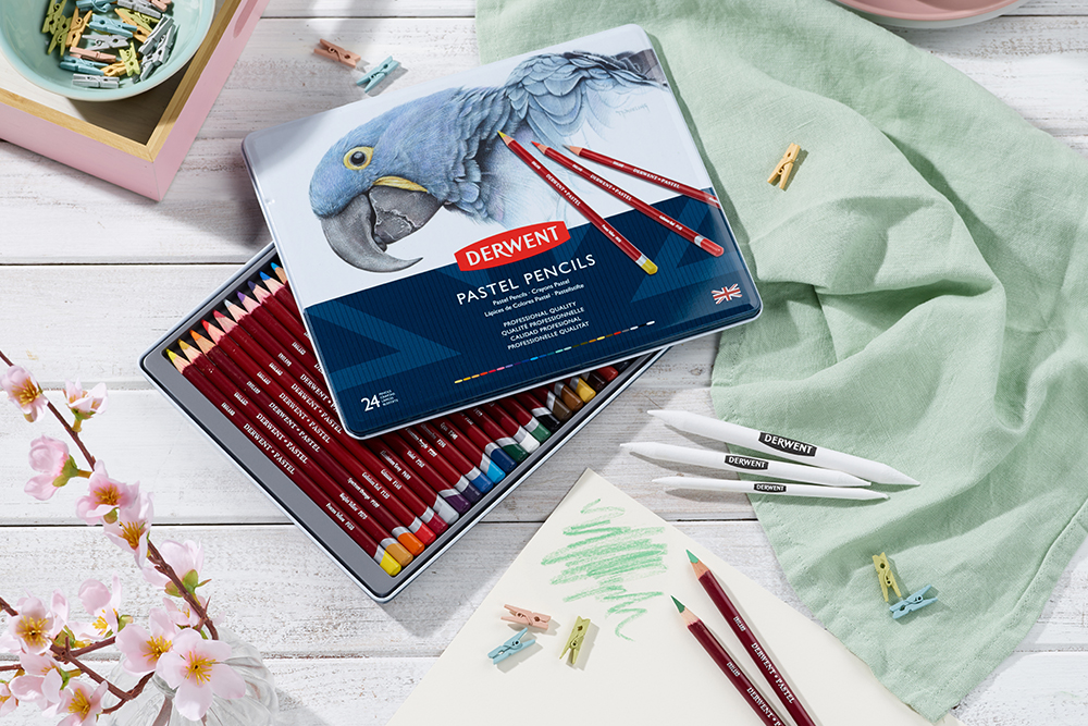 An open tin of 24 Derwent Pastel Pencils. The tin is photographed on a white wooden background showing the contents of the tin. Two pastel pencils are loose and have been used to create colour swatches on a sheet of paper.