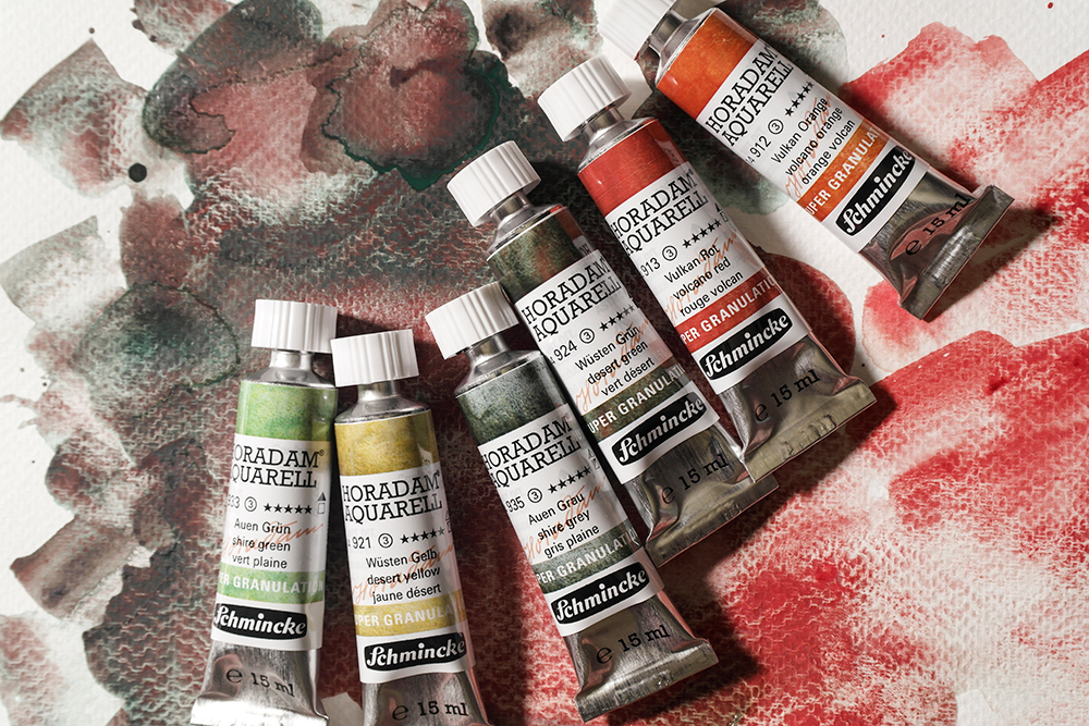 6 15ml tubes of Schmincke Horadam Aquarell Super Granulating Watercolour Paint in Shire Green, Desert Yellow, Shire Grey, Desert Green, Volcano Red and Volcano Orange on a background wash of Desert Green and Volcano Red.