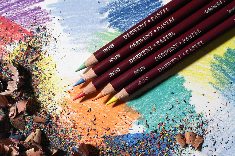 Assorted Derwent Pastel Pencils are photographed on a background covered in pastel marks and pencil sharpenings.