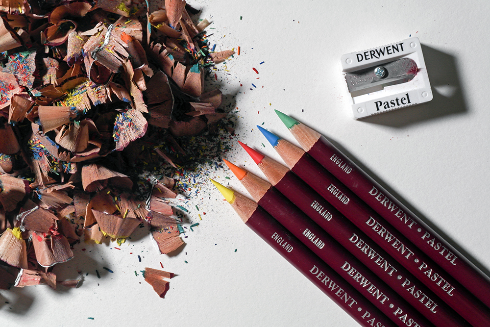 Keep Your Pastel Pencils Sharp - 5 Derwent Pastel Pencils in Yellow, Oraneg, Red, Blue and Green are photographed with a pastel pencil sharpener next to some pencil shavings.