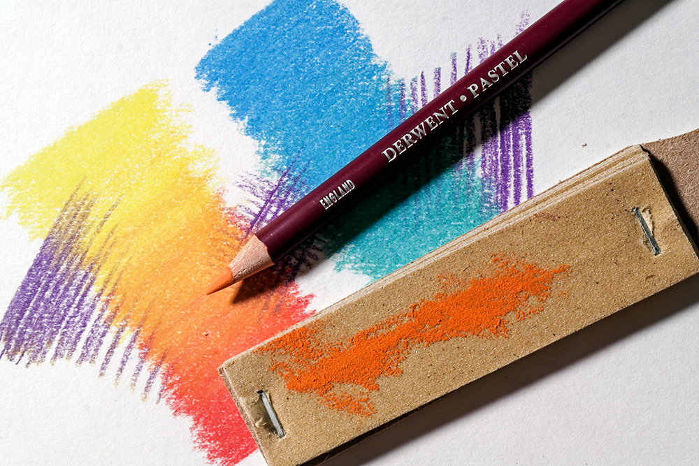 An orange Derwent Pastel Pencil and Loxley Sandpaper Block are photographed on a colourful pastel background. The sandpaper block has been used to hone the tip of the pencil.