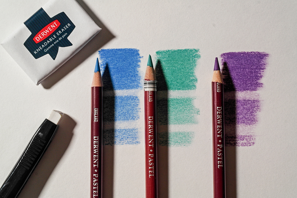 Lift pastel to create interesting highlights - three Derwent Pastel Pencils in Turquoise, Blue and Purple are photographed next to swatches. The swatches have been erased in areas with a rubber.