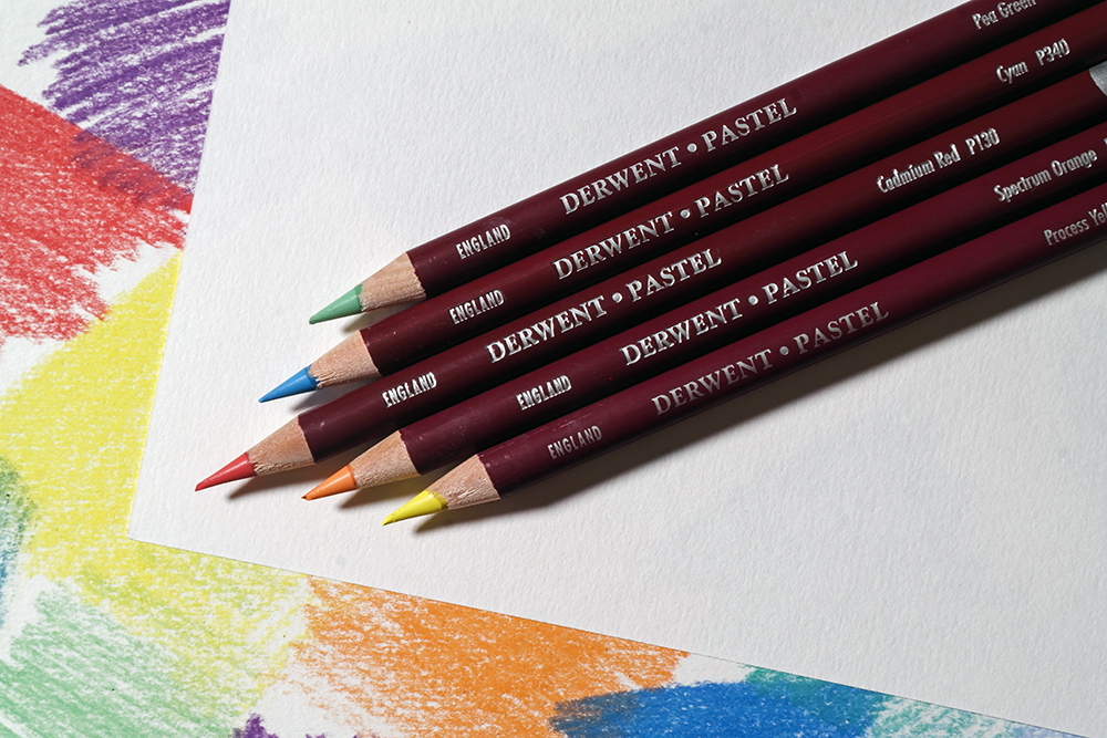 Protect your work with a sheet of clean paper - Derwent Pastel Pencils are photographed on a clean paper sheet, on top of paper covered in soft pastel marks.