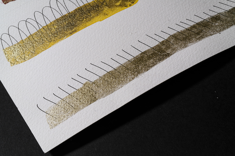 Two swatches on Aqua Bronze applied over black fine line pen. One swatch is the Aqua Bronze on it's own, in the second swatch the Aqua Bronze has been mixed into yellow watercolour paint.