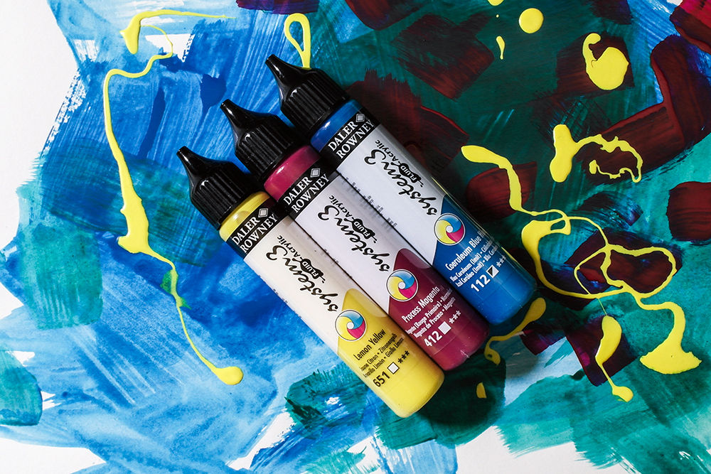Three bottles of System 3 Fluid Acrylic Paint in Lemon Yellow, Process Magenta and Coeruleum Blue Hue on a painted background