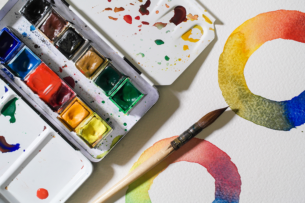 A metal box containing half pan watercolours rests on a sheet painted with two colour wheels.