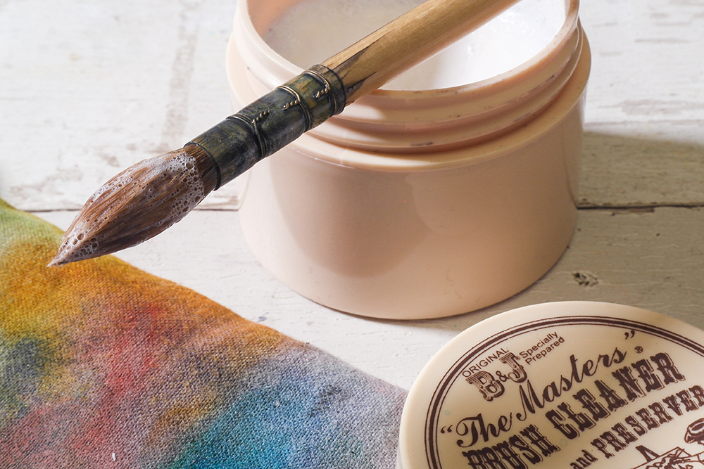 A quill shaped watercolour brush is photographed resting on a jar of Masters Brush Cleaner.