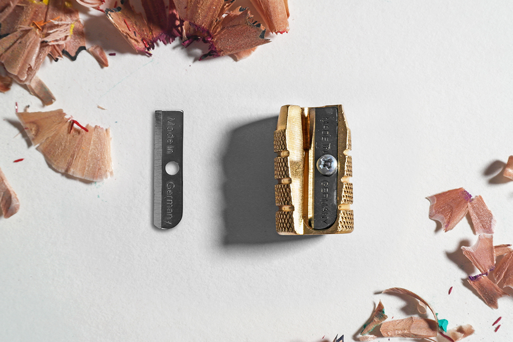 A single loose sharpener blade photographed next to brass bullet pencil sharpener, on a white background.