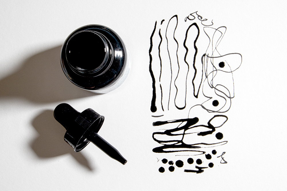 A bottle of Winsor & Newton Black Indian Drawing Ink and glass pipette dropper lid alonside scribbled lines drawn with the pipette.