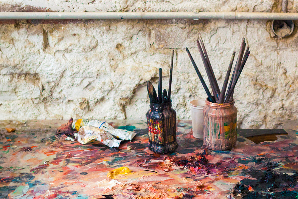 Used artists paint brushes sit in jars on a messy artists workbench